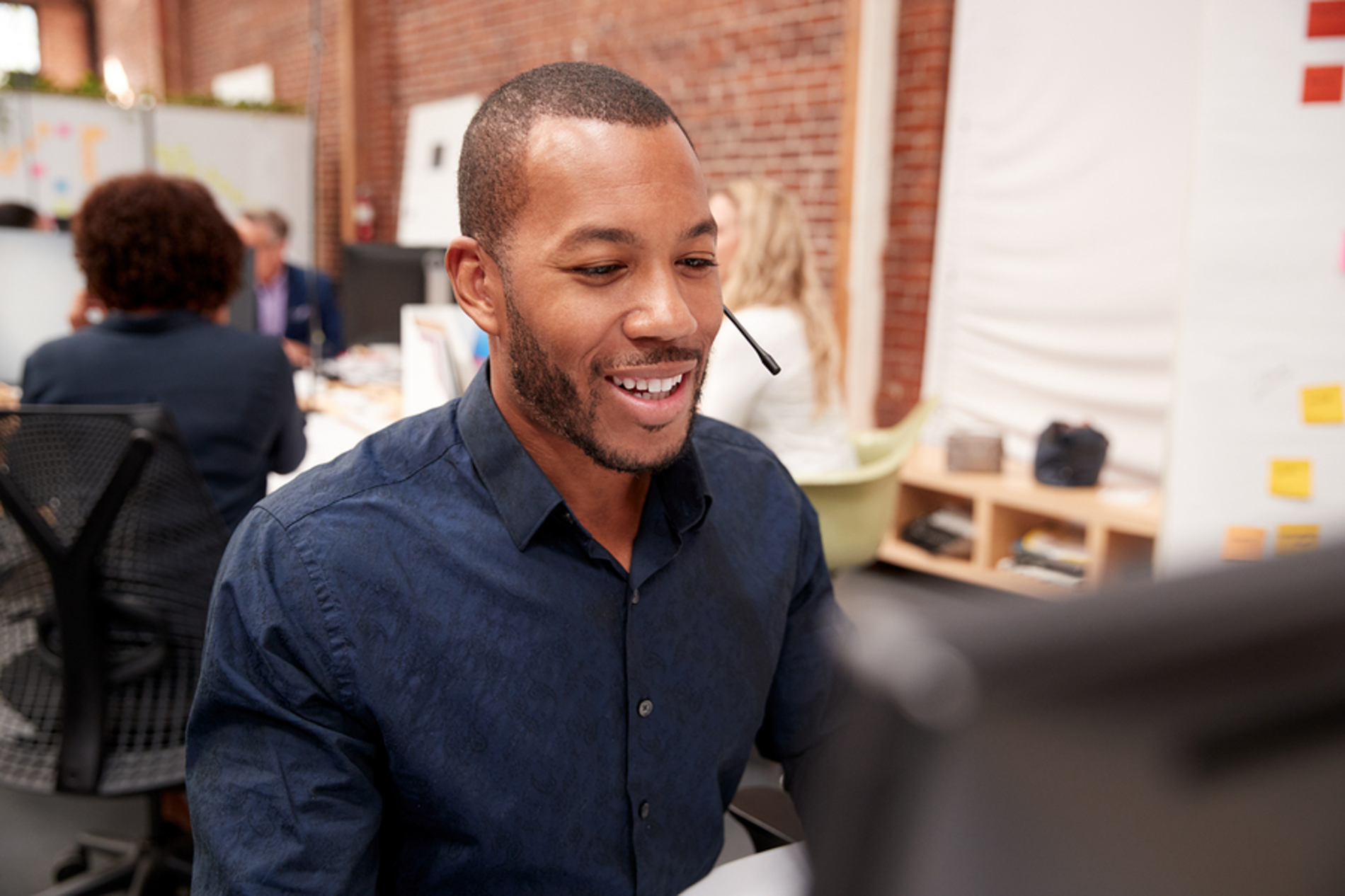 Smiling man in front of a desk top computer speaking into a headset.